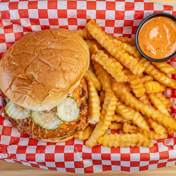 Kluckin’ Chicken sandwich with pickles, crinkle-cut fries, and dipping sauce in a red checkered basket.