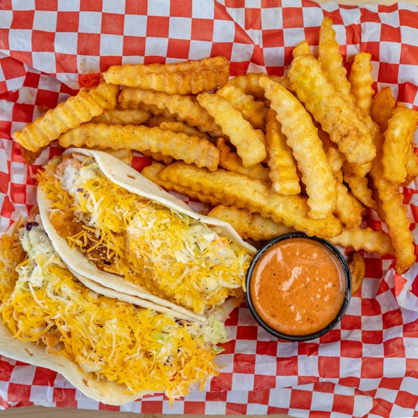 Two Kluckin' Chicken tacos with shredded cheese and coleslaw, crinkle-cut fries, and dipping sauce.