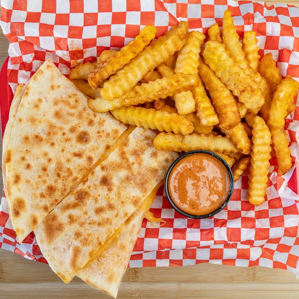 Quesadilla slices, crinkle-cut fries, and a dipping sauce in a red and white checkered basket.