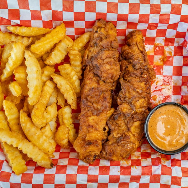 Two crispy fried chicken tenders, crinkle-cut fries, and a side of dipping sauce on checkered paper.